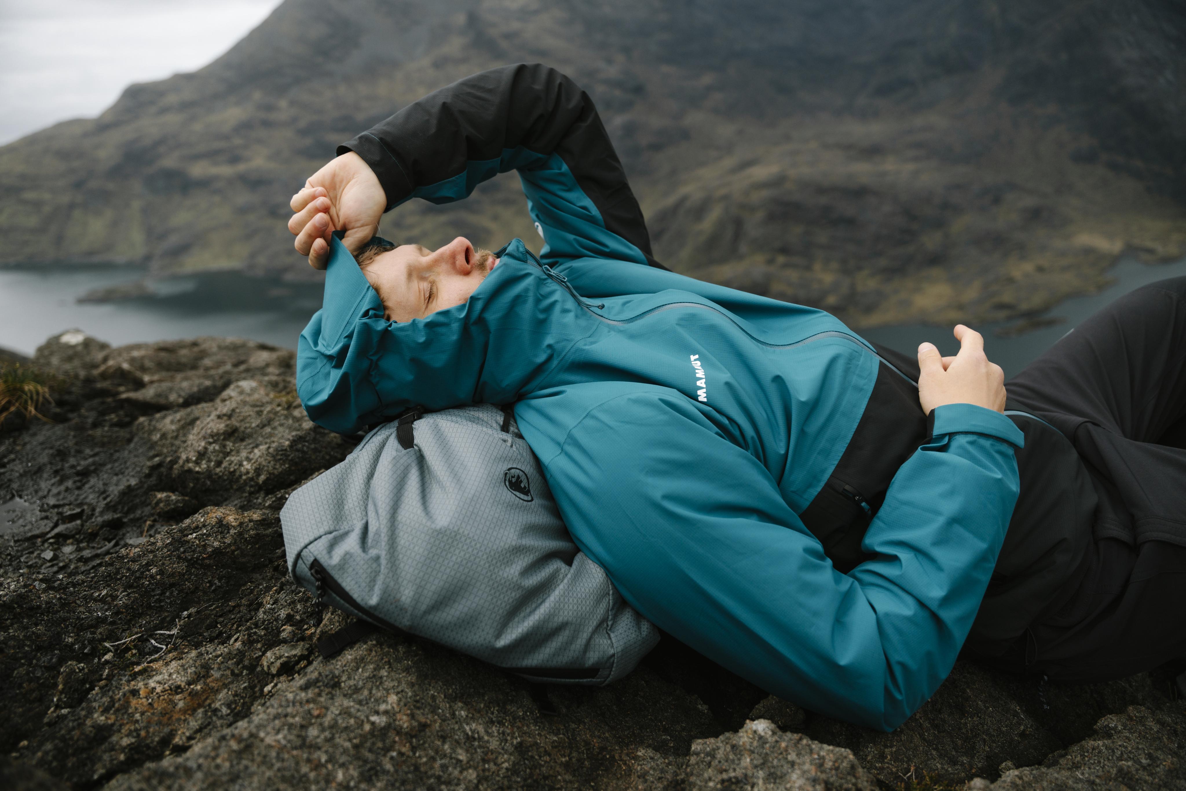 A man wearing Mammut sportswear and a cap stretches outdoors on a mountain trail, with water bottles attached to his running vest.